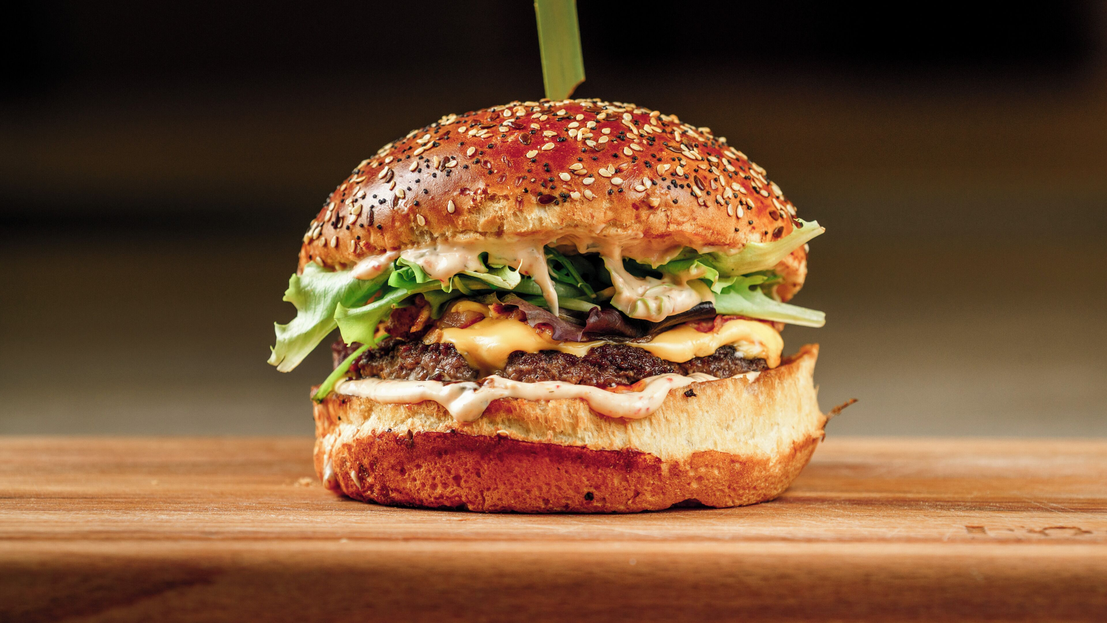 A burger with sesame seed bun, lettuce, cheese, and sauce, placed on a wooden surface.
