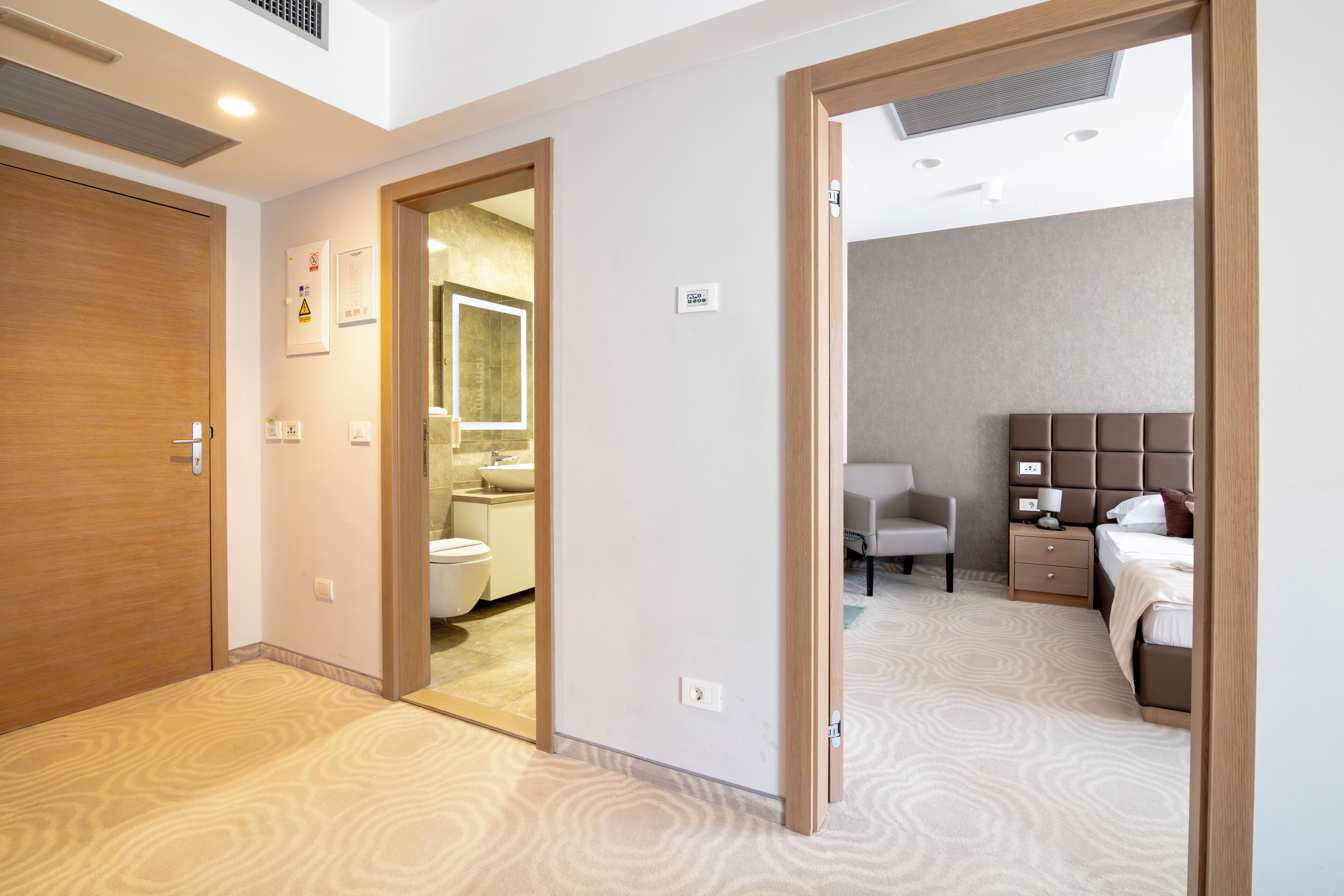A hotel room with a wooden door, a bathroom with a toilet and sink, a bed with a headboard, and a chair.