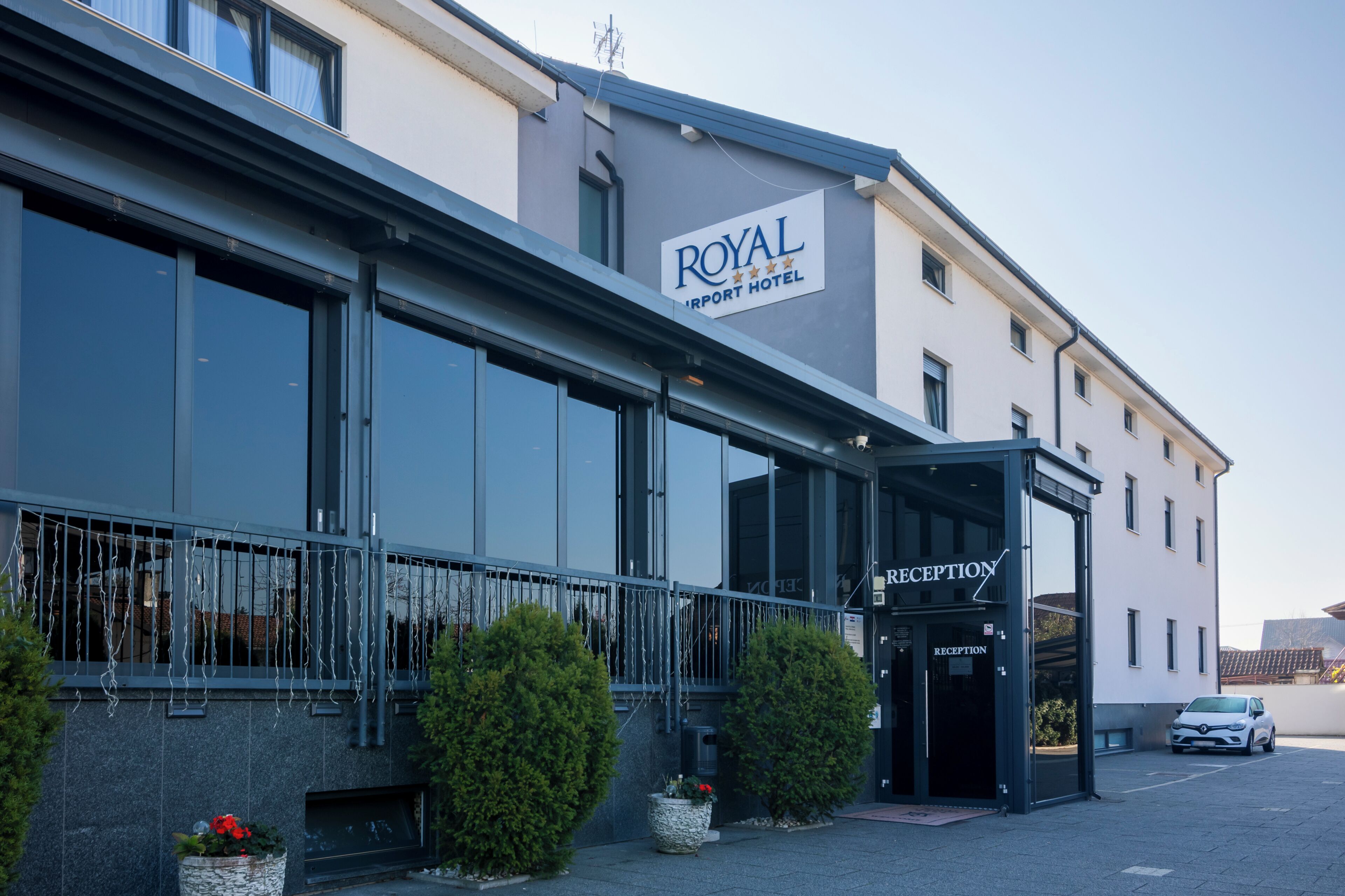 Een hotelingang met een receptiebalie, een bord met de tekst 'ROYAL Airport Hotel' en een geparkeerde auto.