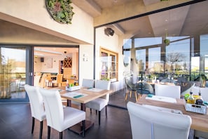 Un comedor moderno con sillas y mesas blancas, un separador de vidrio y vistas al exterior.