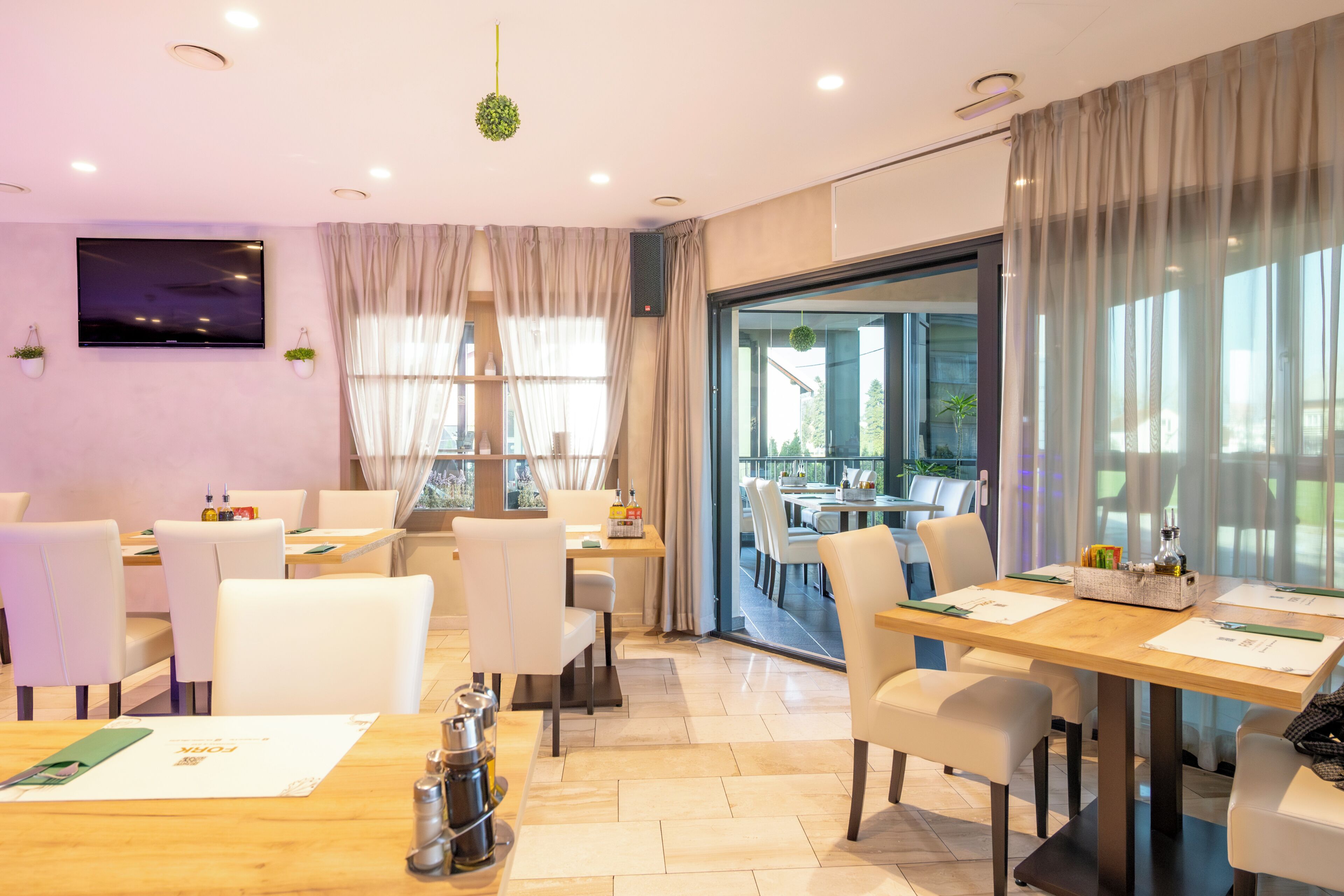 A modern dining area with tables and chairs, a flat-screen TV on the wall, and large windows with curtains.