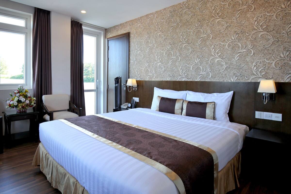 A hotel room with a large bed, a desk with a lamp, a chair, and a window with curtains.