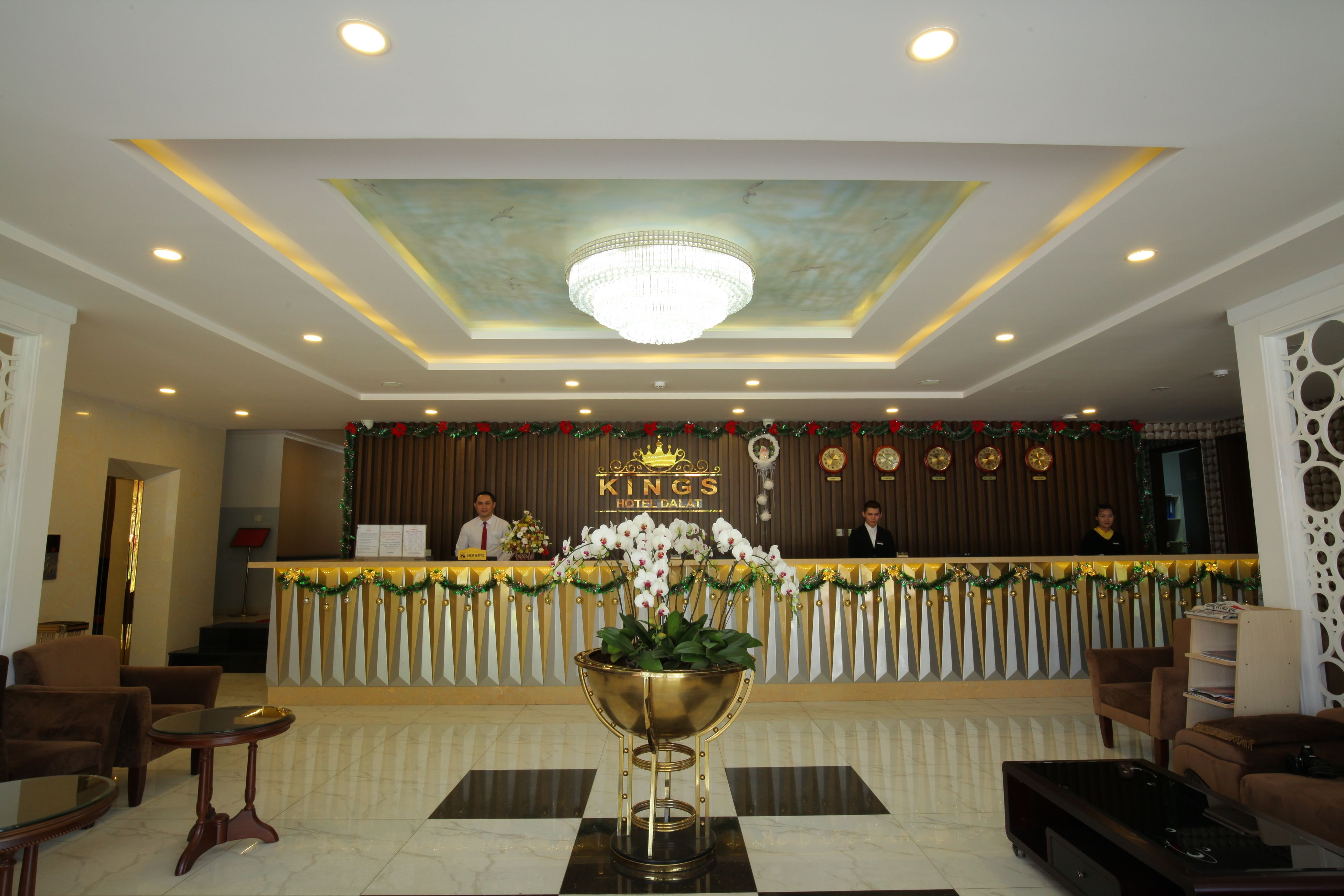 Zona de recepción de un hotel con un mostrador dorado, una araña de cristal y un arreglo floral grande.