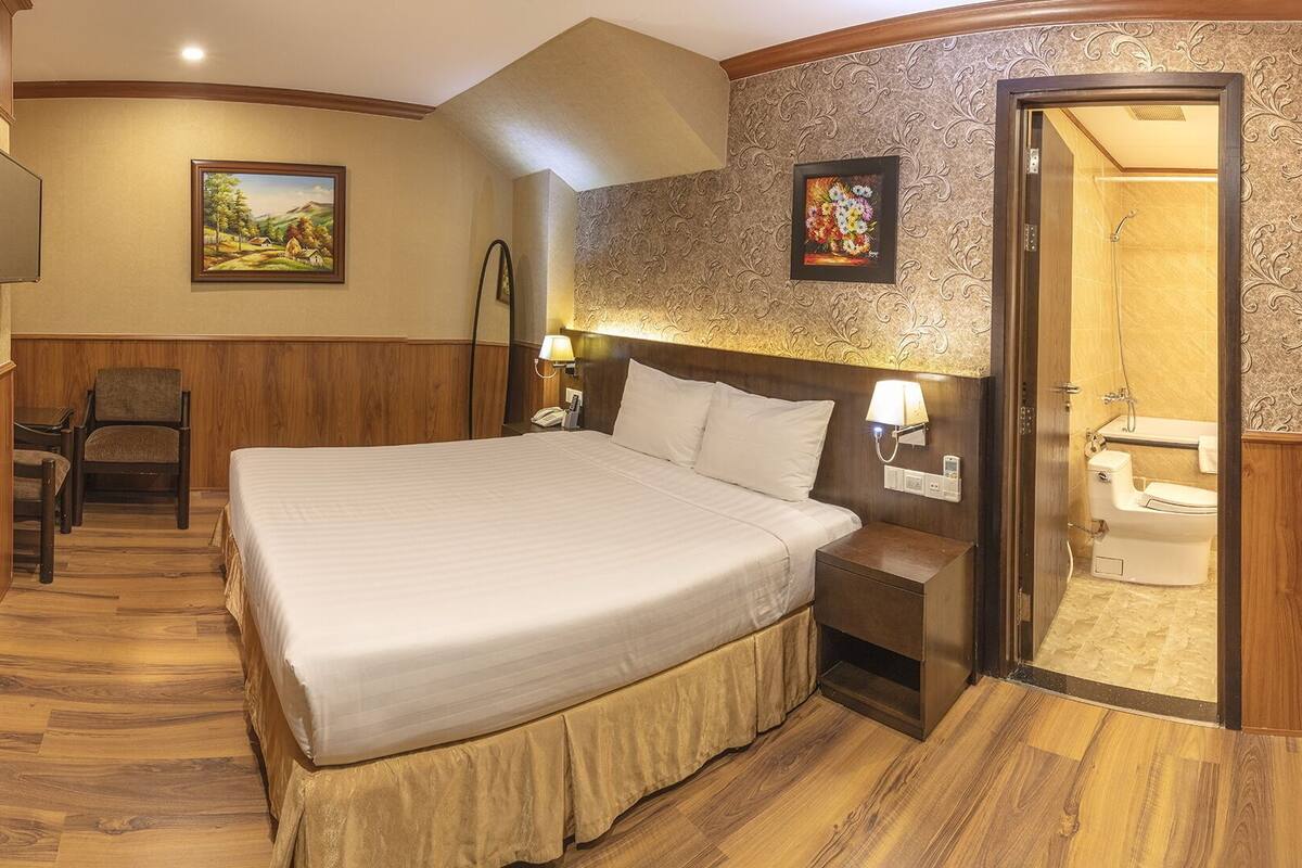 A hotel room with a large bed, a desk with a chair, a telephone, and a painting on the wall.