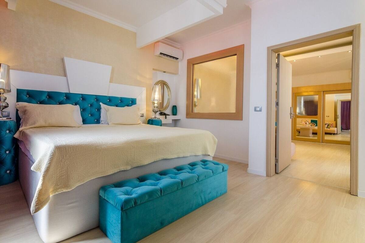 A bedroom with a large bed, a bedside table, a mirror, and a door leading to another room.