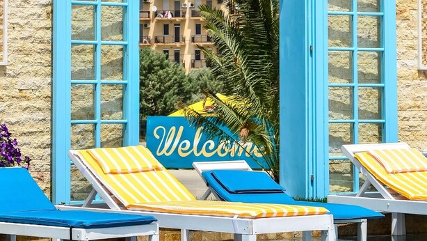 Zona de piscina con puertas de marco azul, sillas reclinables a rayas amarillas y un cartel que dice 'Welcome'.