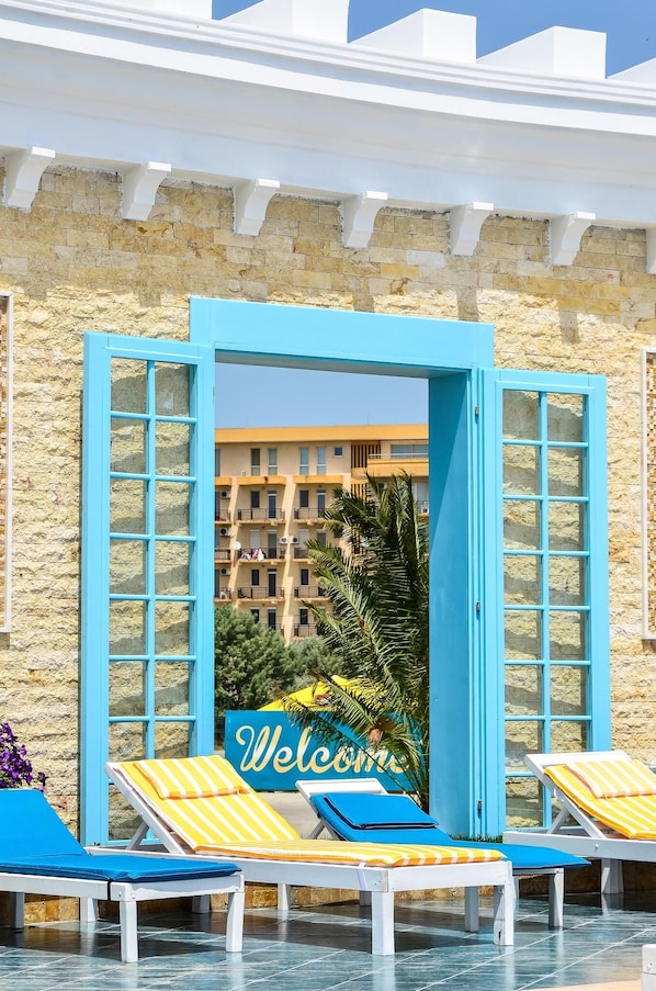 Zona de piscina con puertas de marco azul, sillas reclinables a rayas amarillas y un cartel que dice 'Welcome'.