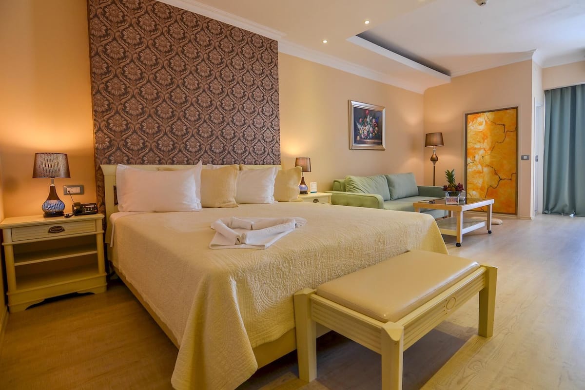 A hotel room with a large bed, a bedside table with a lamp, a green sofa, a small table with a lamp, and a painting on the wall.