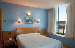 A hotel room with a bed, bedside lamps, a nightstand, a window with curtains, and a view of the outdoors. - Soraya (Suances)