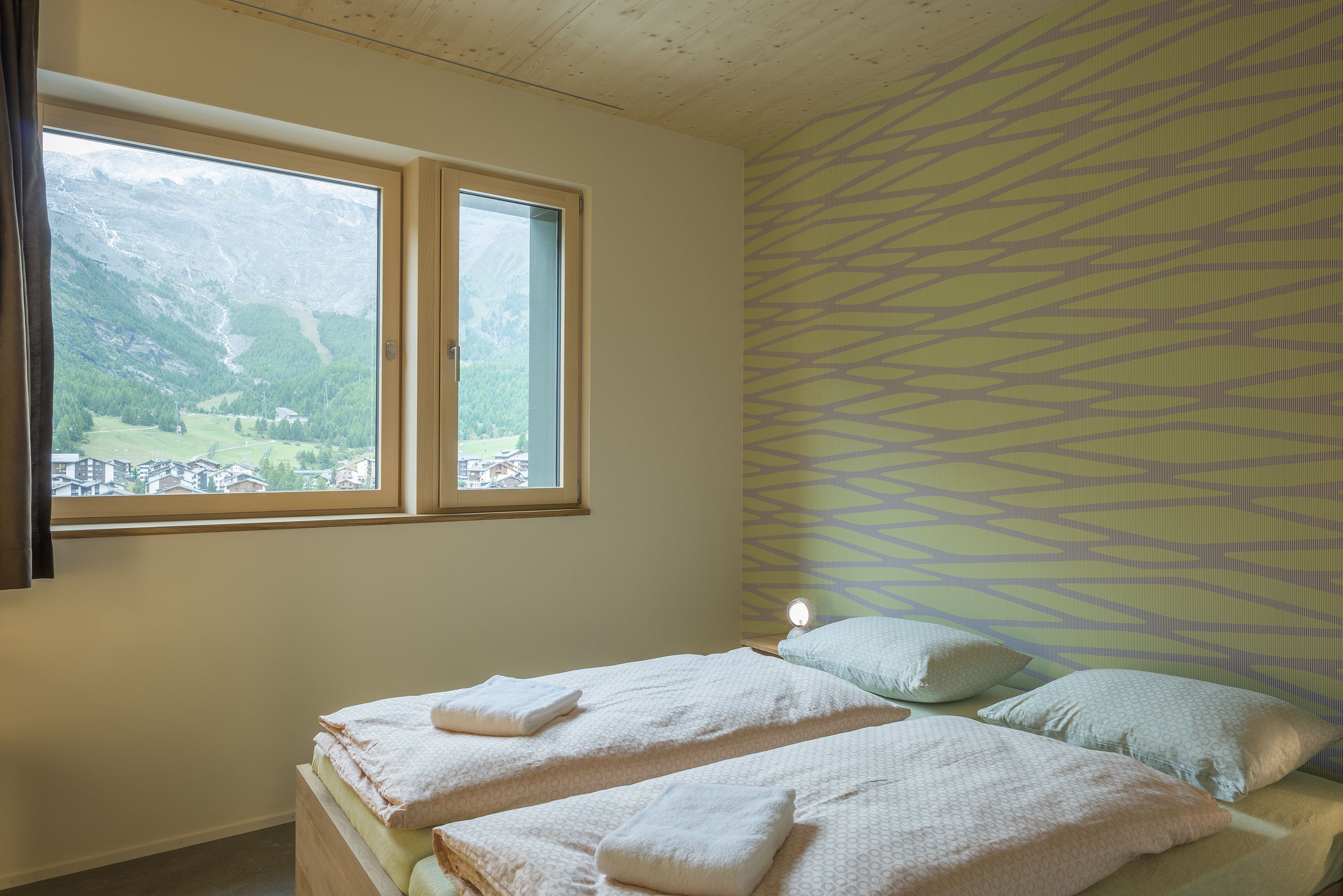 Une chambre avec deux lits, une fenêtre donnant sur la montagne et un mur décoré d’un motif.