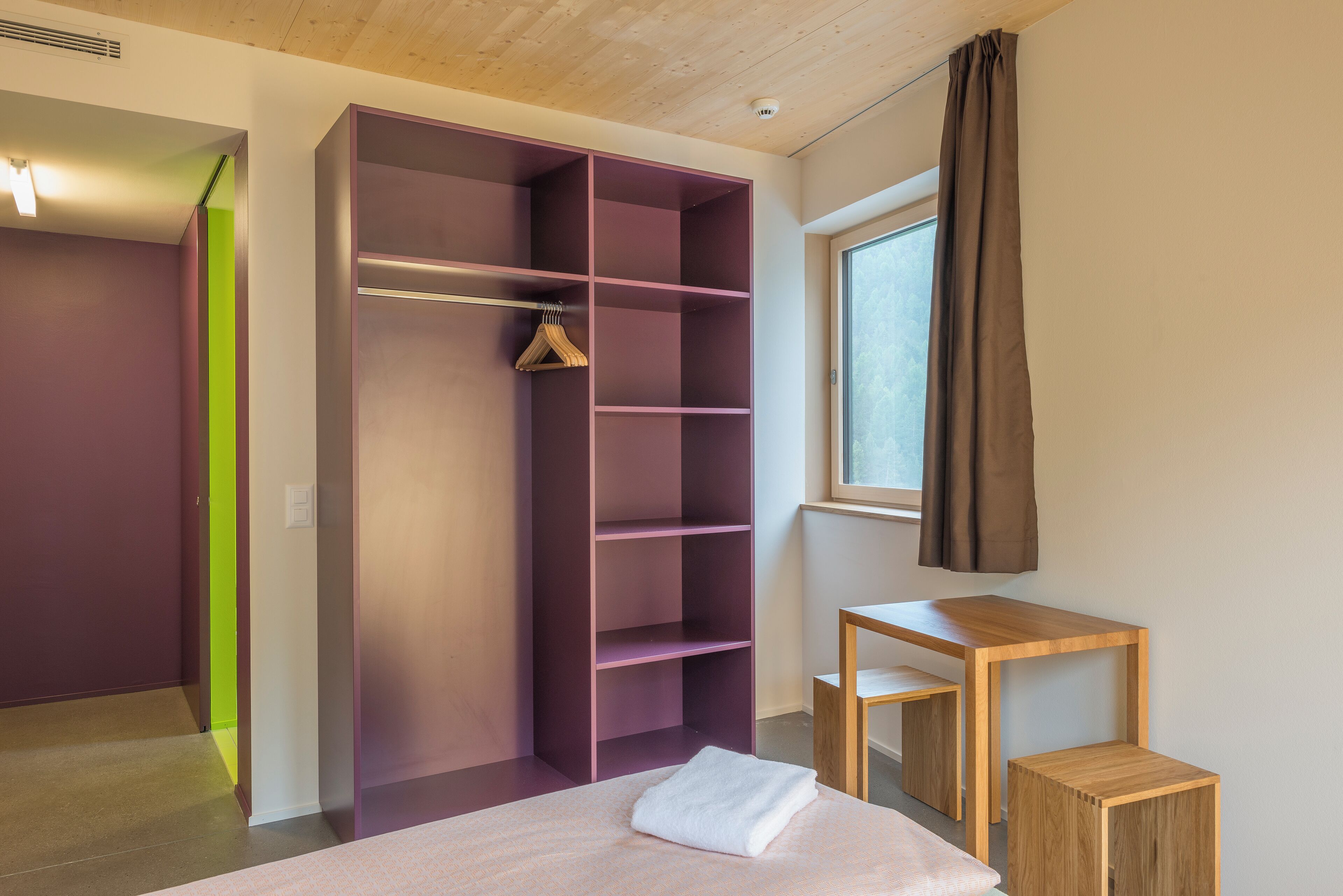 A bedroom with a bed, a wooden nightstand, a purple wardrobe, and a window with curtains.