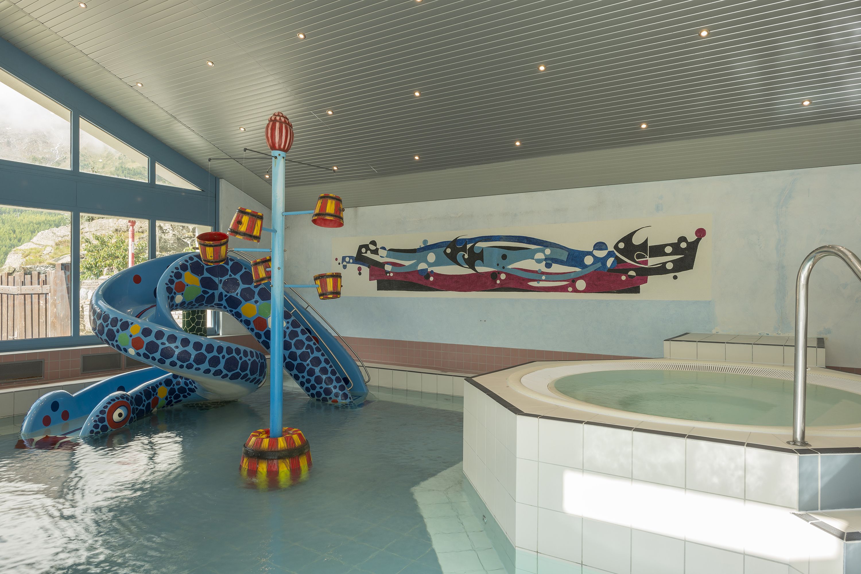 Een binnenzwembad met een blauwe glijbaan, een jacuzzi en een muurschildering.