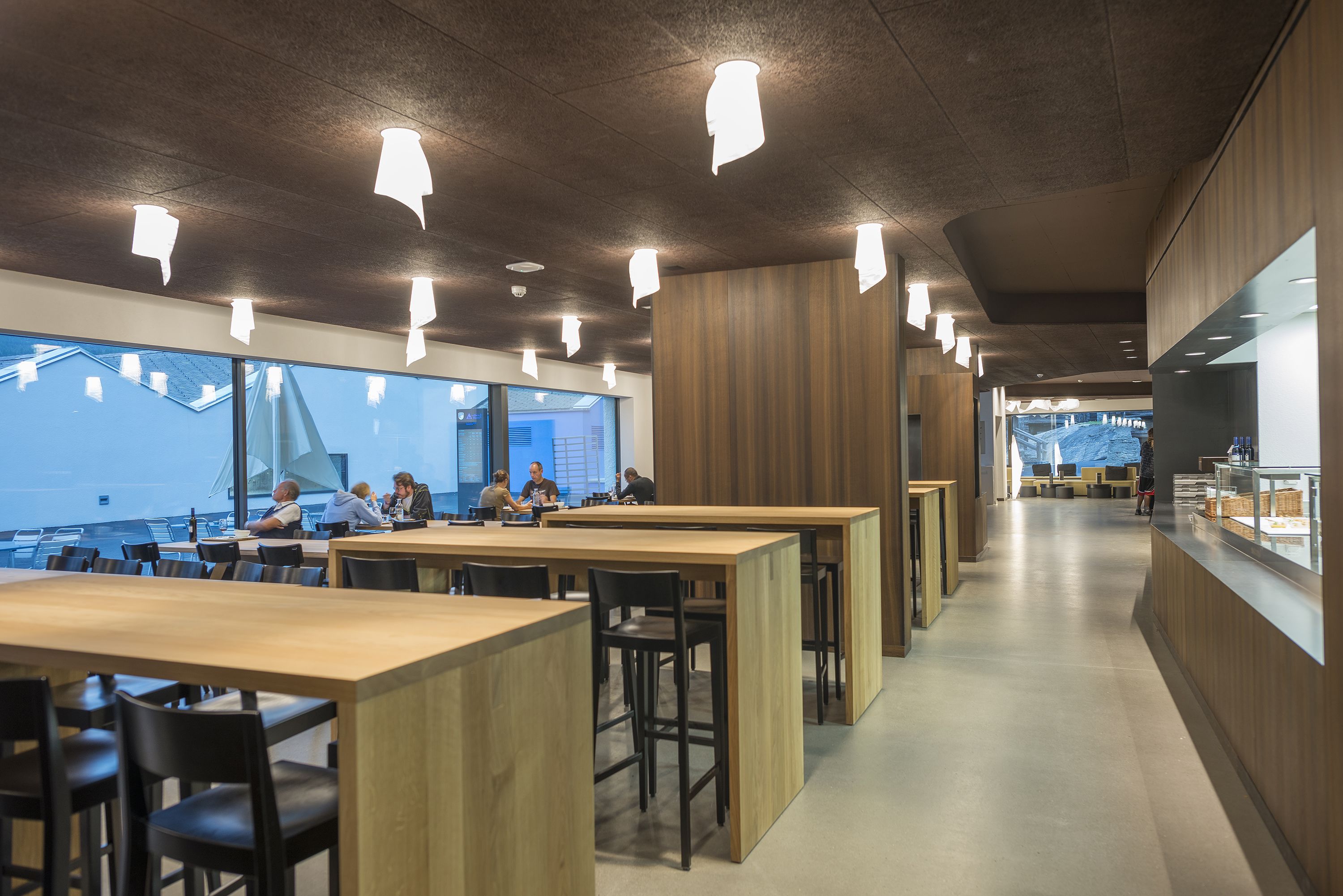 A modern dining area with long wooden tables and black chairs, large windows offering a view of the sea, and pendant lighting.