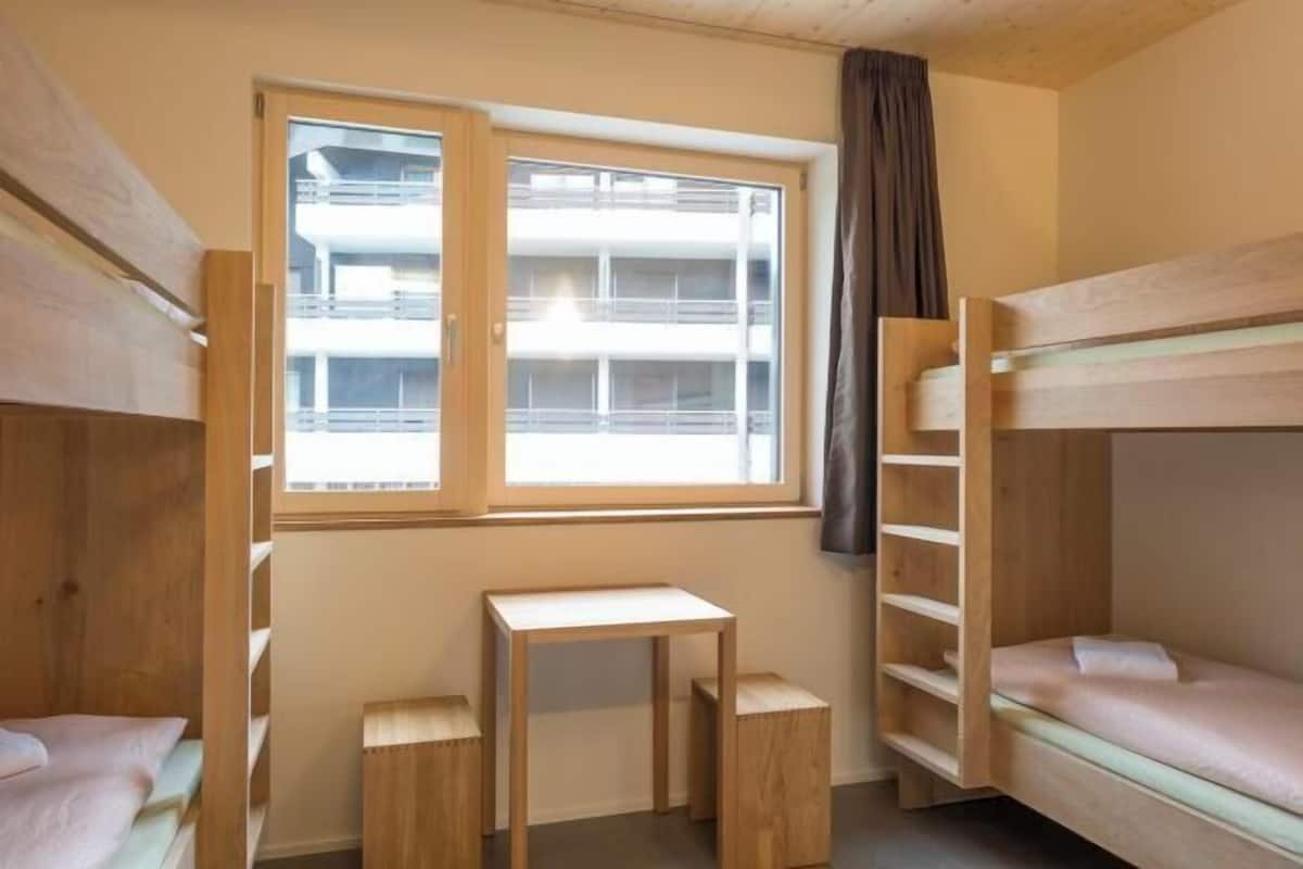 A bunk bed room with a window, a small table, and two stools.