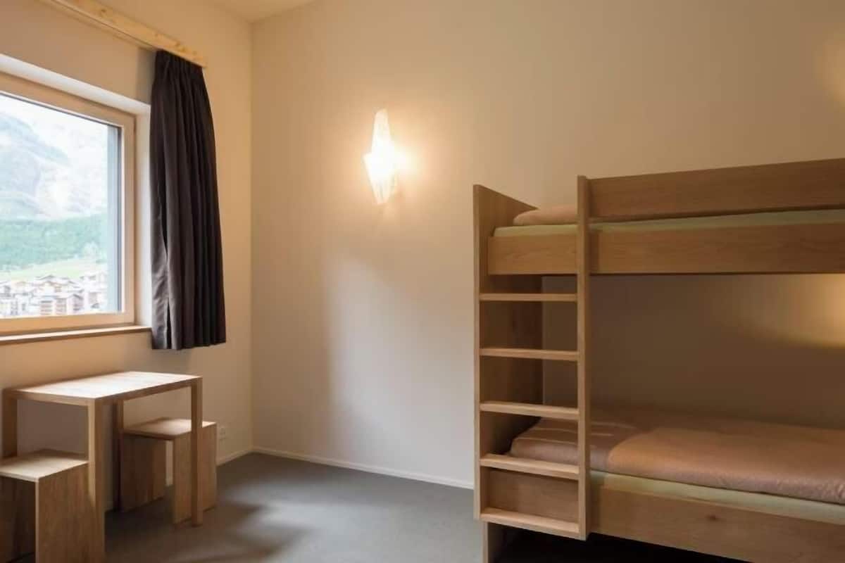 A bunk bed with a view of a mountainous landscape through a window, a desk, and a wall-mounted light fixture.