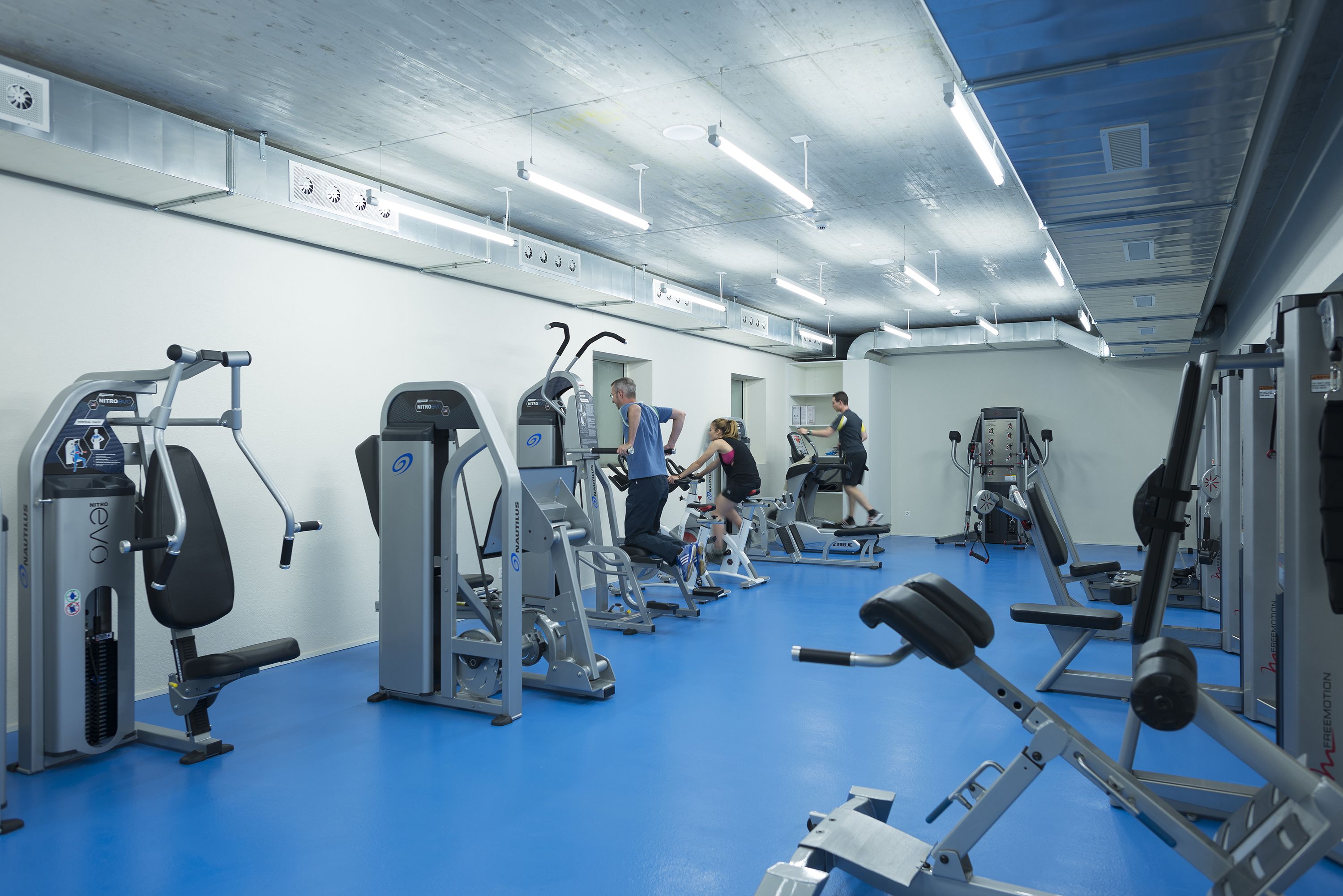 Un gimnasio con diversas máquinas de ejercicio, suelo azul y paredes blancas.
