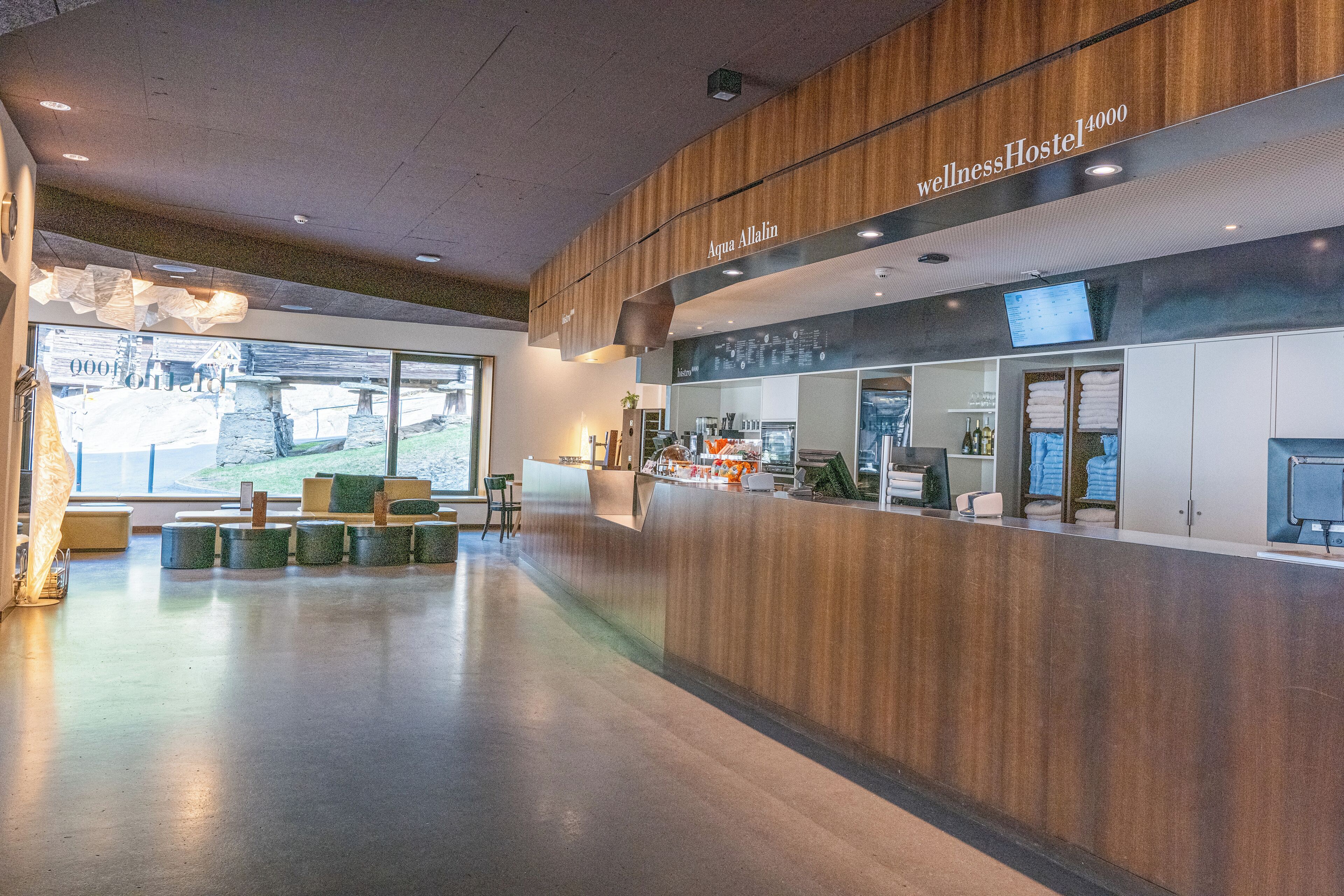 A modern wellness hostel interior with a reception desk, seating area, and a large window offering a view of the outdoors.
