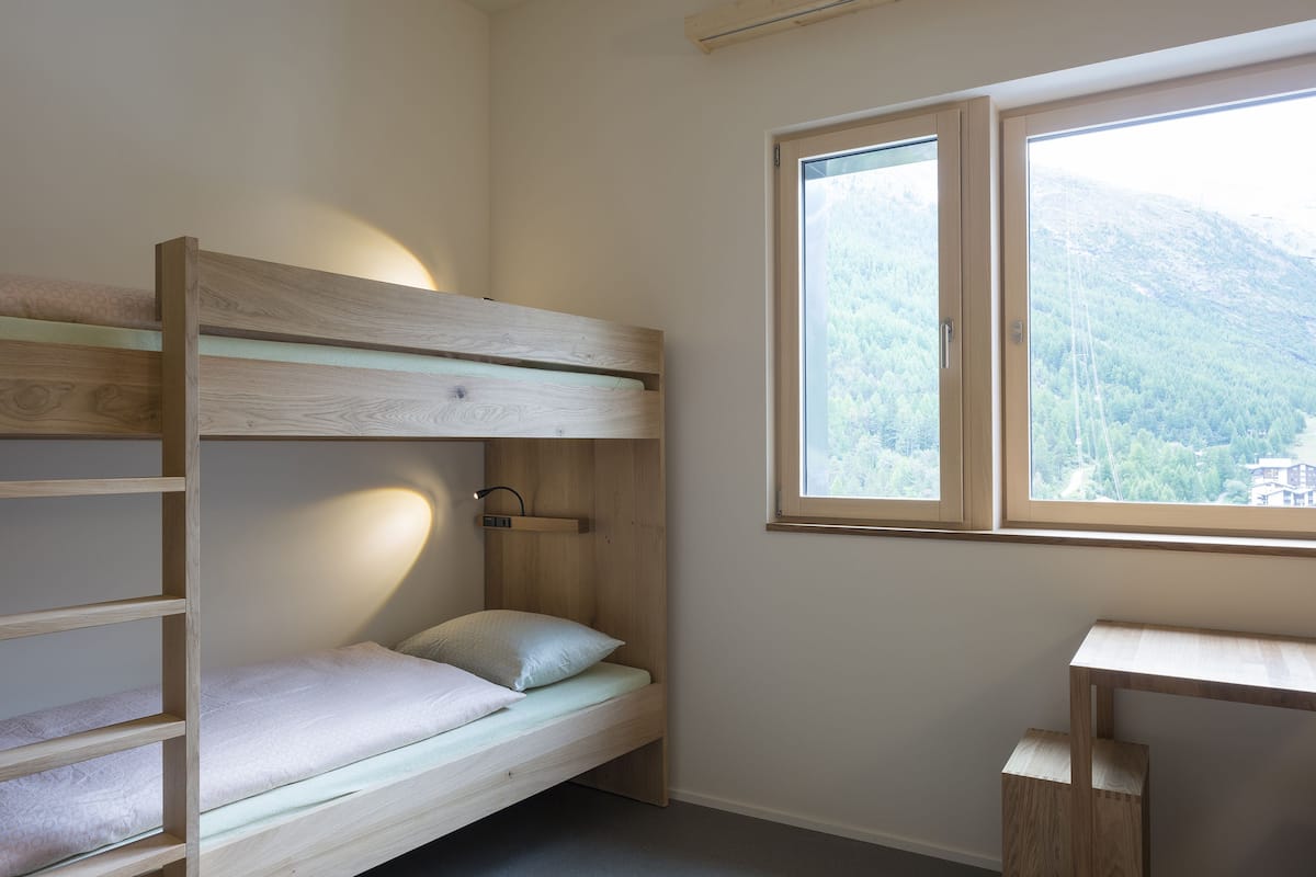 A bunk bed with a wooden frame, a bedside table, and a window with a view of a forested mountain.