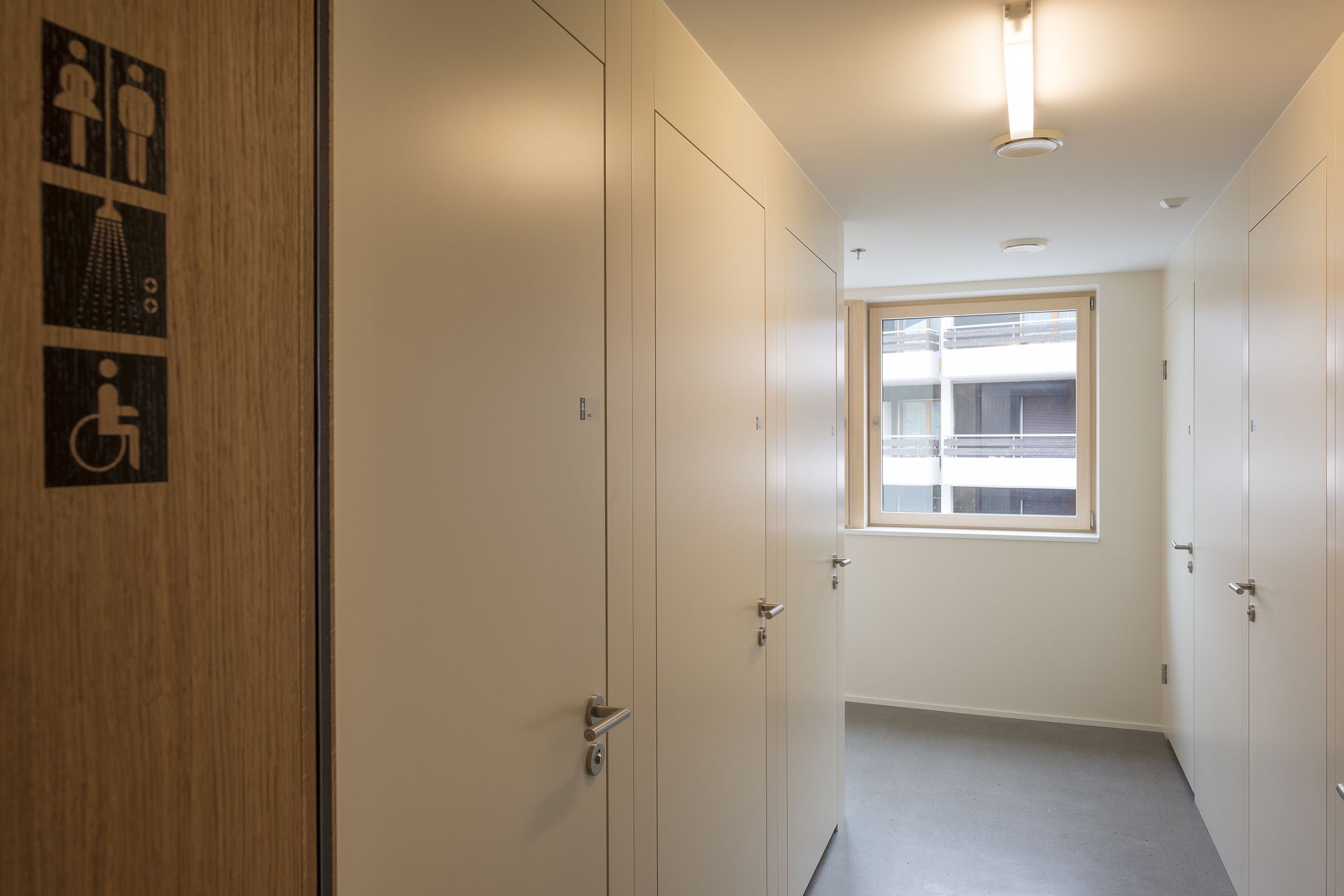 Passadís amb una porta de fusta a l'esquerra, una finestra al final i una porta amb el símbol de cadira de rodes.