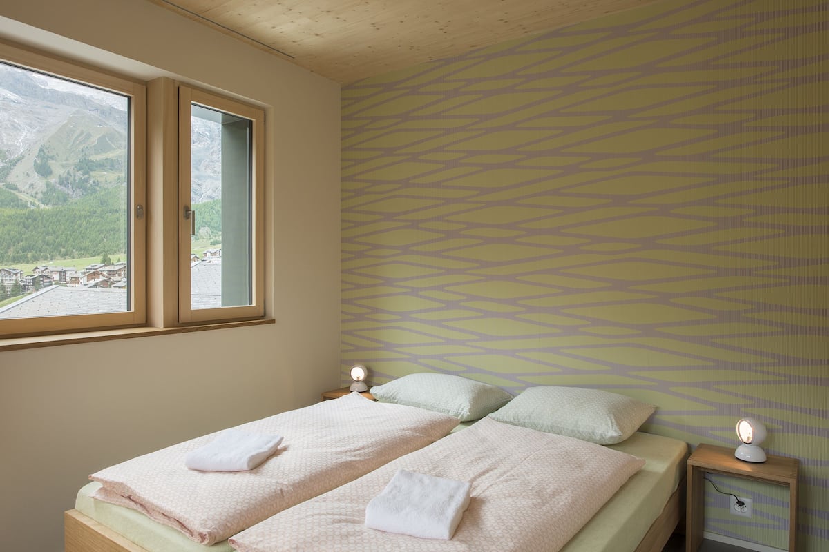 A bedroom with two beds, a window with a view of mountains, and a patterned wall.