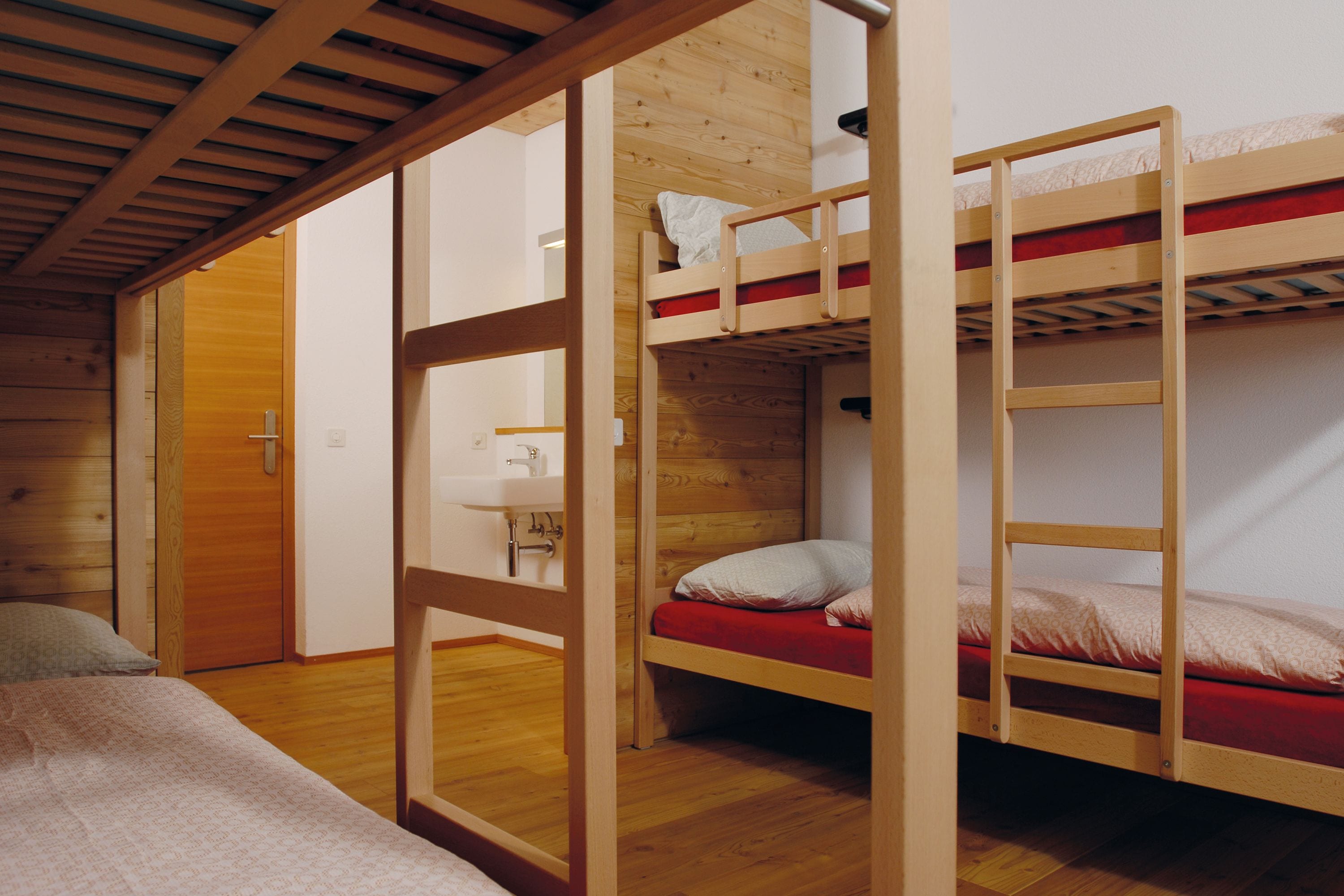 Une chambre avec deux lits superposés, un plancher en bois, une porte et une petite table de chevet avec une lampe.
