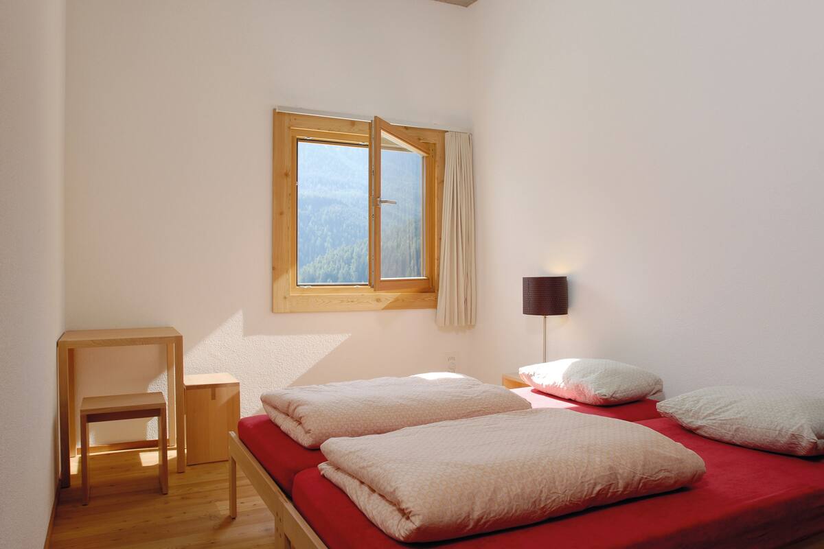 A room with a bed, a wooden table, a lamp, and a window with a view of trees.