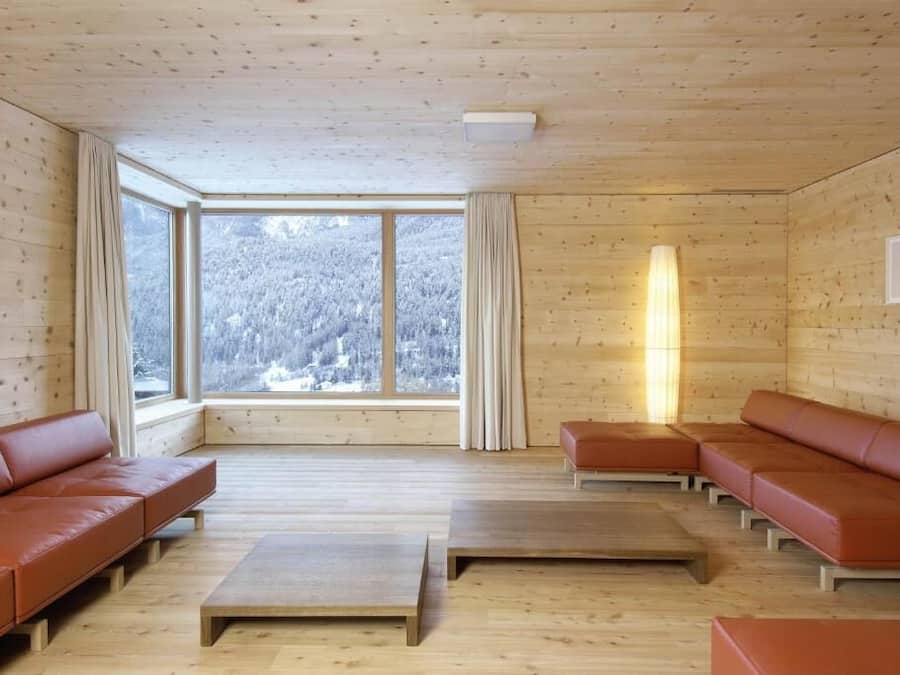 A spacious room with wooden walls and floors, large windows offering a view of a snowy forest, and a seating area with leather sofas and wooden tables.