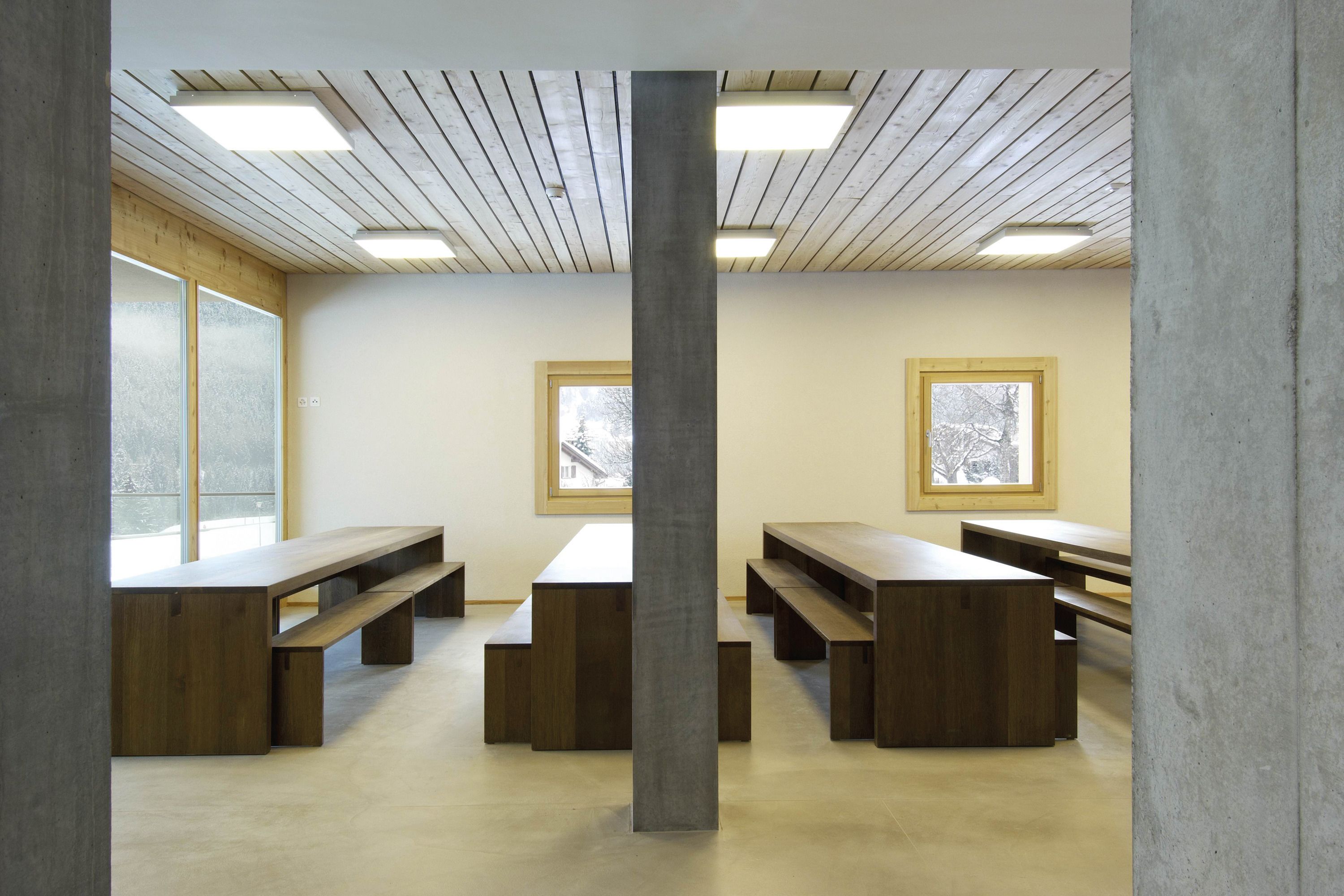 Una habitación con bancos de madera y un pilar central, ventanas amplias y un techo inclinado con luces.