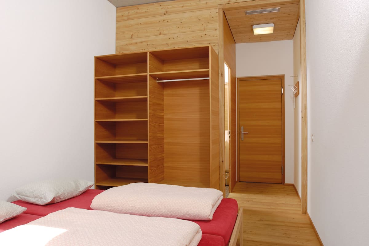 A bedroom with a large wooden wardrobe, a bed with red bedding, and a wooden floor.
