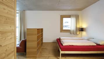 Una habitación con piso de madera, una cama con colchón rojo, un armario de madera, una silla roja y una ventana con cortinas.