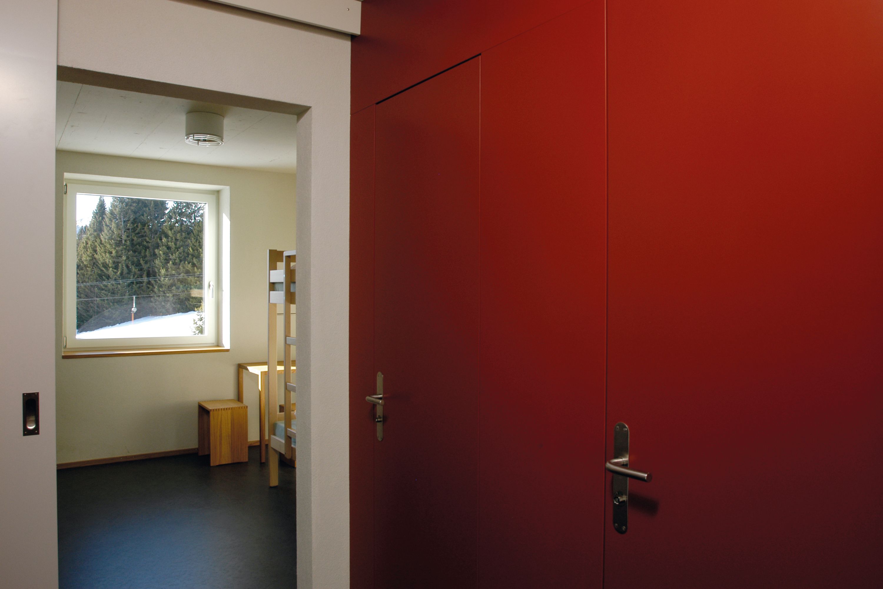 A room with a red door, a window with a view of trees, and a small wooden stool.