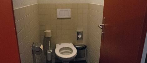 A small, narrow bathroom with a toilet, a trash bin, and a paper towel dispenser.