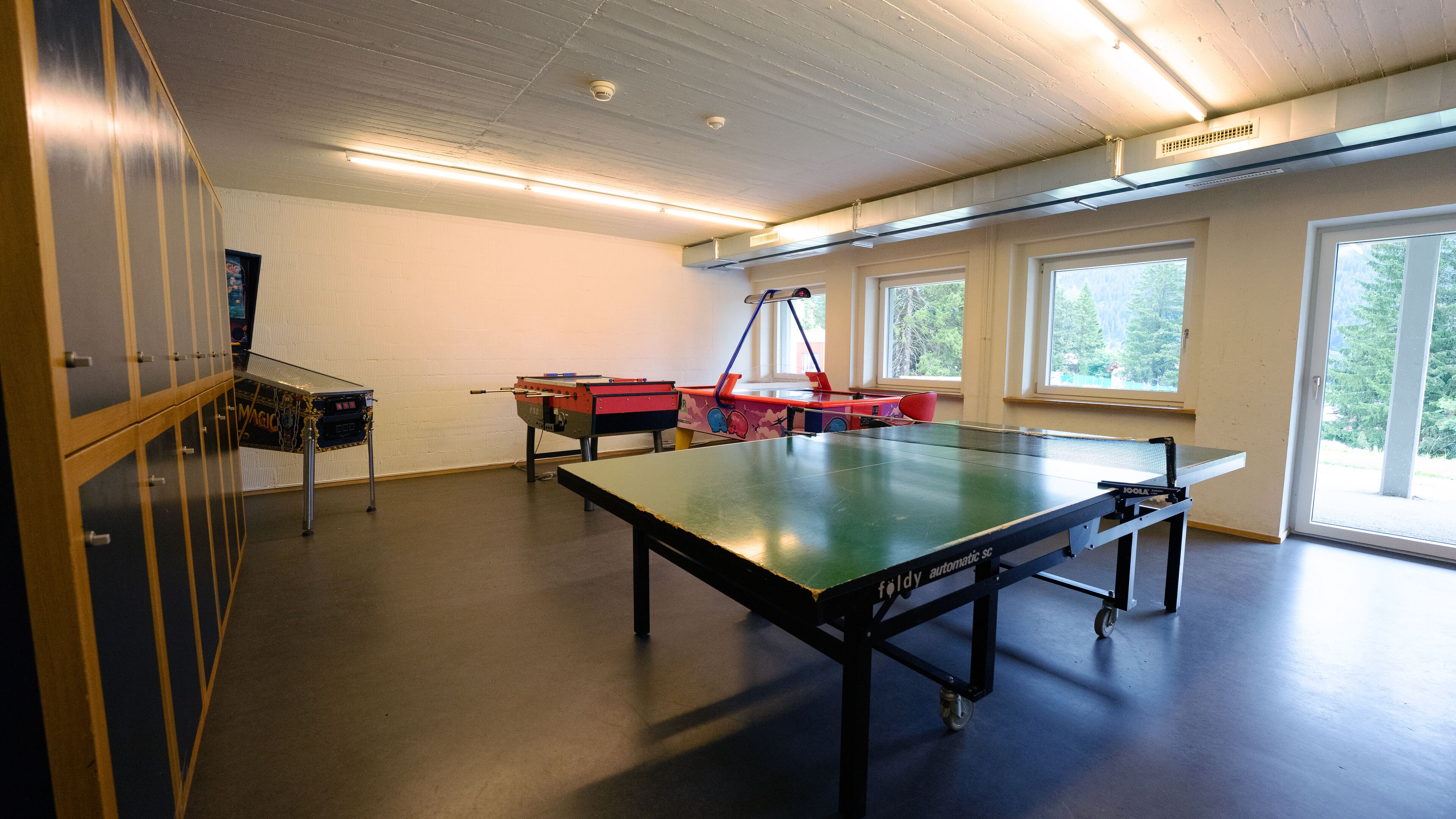 Una habitación con mesa de ping pong, mesa de futbolín y mesa de tenis de mesa. Hay ventanas con vistas a árboles.