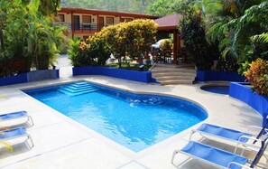 Una piscina con agua azul, rodeada por un deck con azulejos azules y blancos. Hay sillas de descanso y un área de estar cubierta con una mesa y sillas.