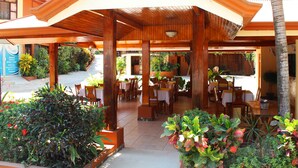 An outdoor restaurant with wooden tables and chairs, surrounded by potted plants and palm trees. - Hotel Alkamar (Cóbano)