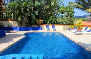 A swimming pool with blue tiles, surrounded by palm trees and lounge chairs. - Hotel Alkamar (Cóbano)