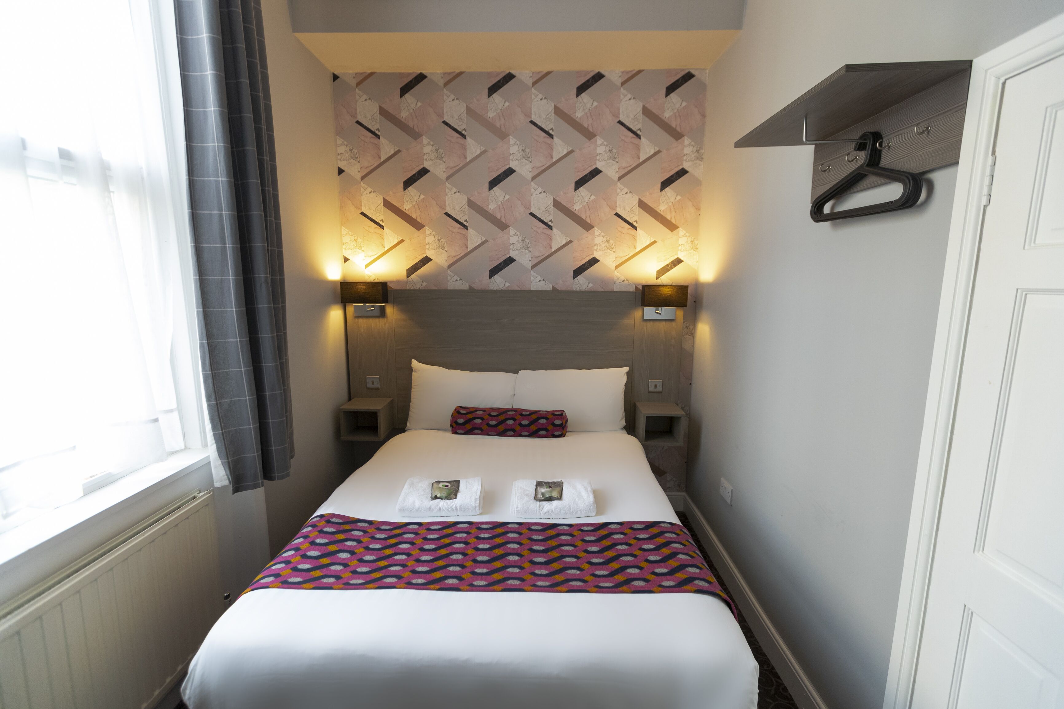 A neatly made bed with a patterned bedspread, two bedside lamps, and a wall-mounted shelf with a hanger.