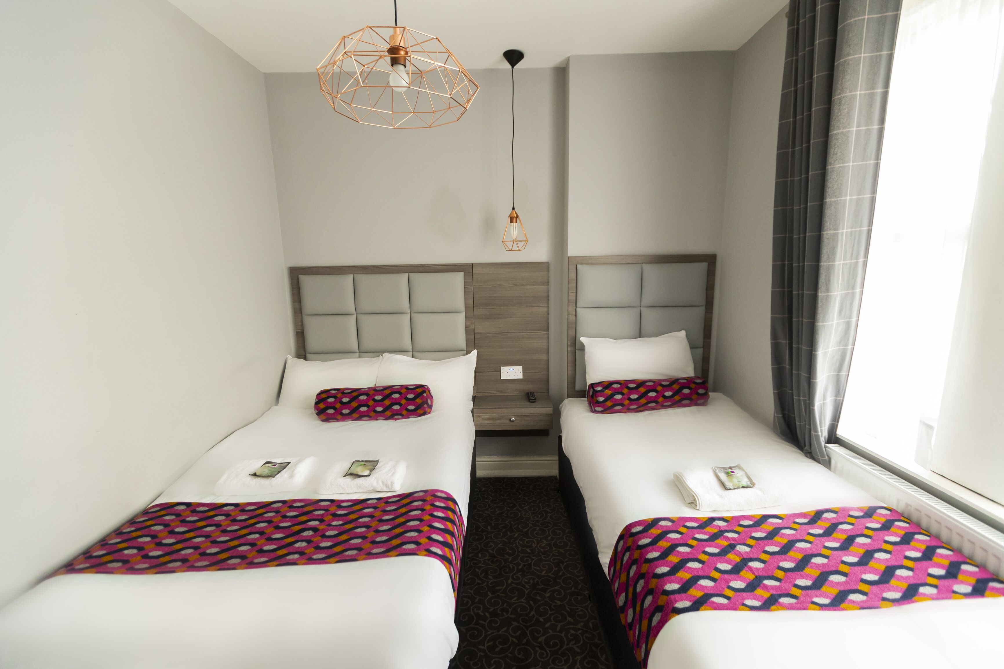 Habitación de hotel con dos camas, cabecero de madera, colcha estampada, una mesita de noche pequeña y una ventana con cortinas.