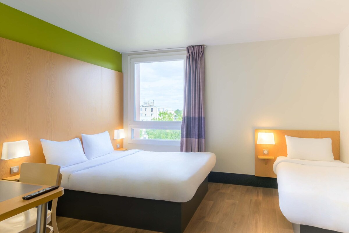 Habitación de hotel con cama, mesitas de noche con lámparas, ventana con cortinas y vistas a edificios.
