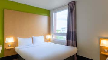 Camera d'albergo con un letto grande, due lampade ai lati del letto, una finestra con tende e vista sugli edifici.