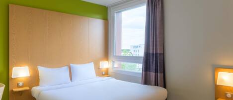 A hotel room with a large bed, two bedside lamps, a window with curtains, and a view of buildings outside.
