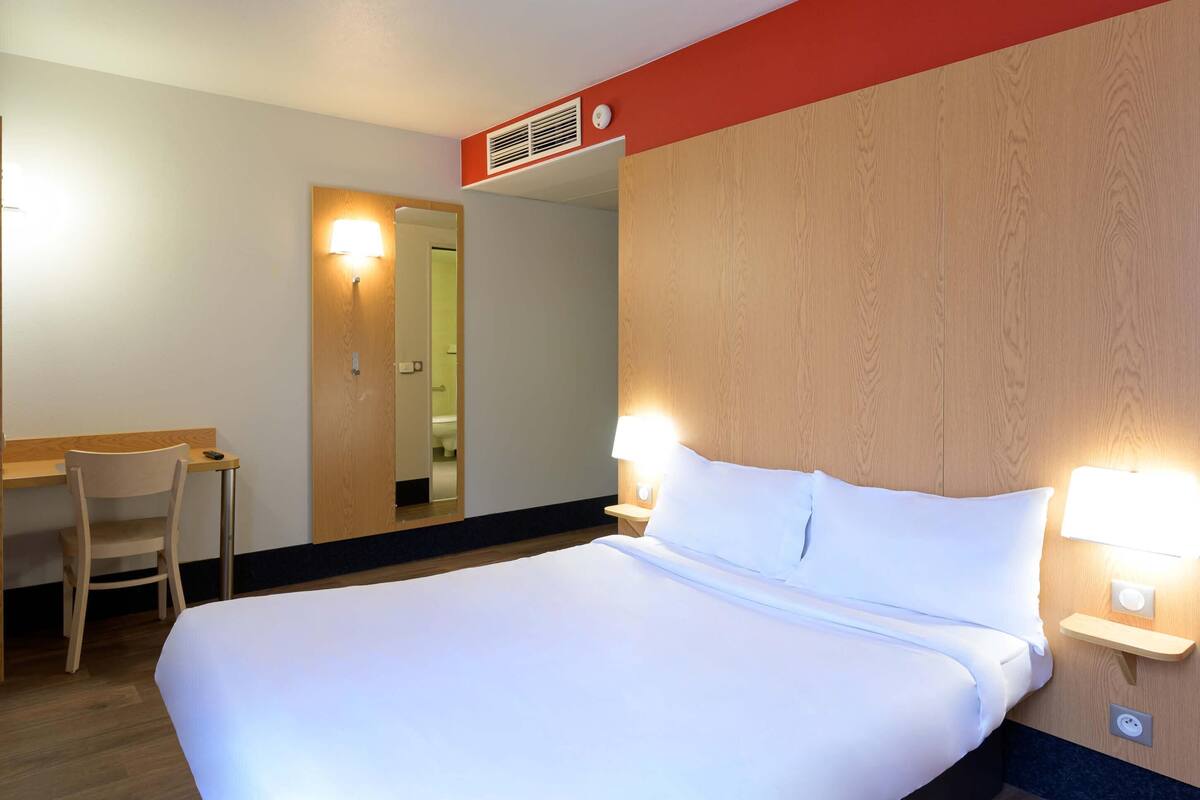 A hotel room with a bed, bedside lamps, a desk with a chair, and a mirror.