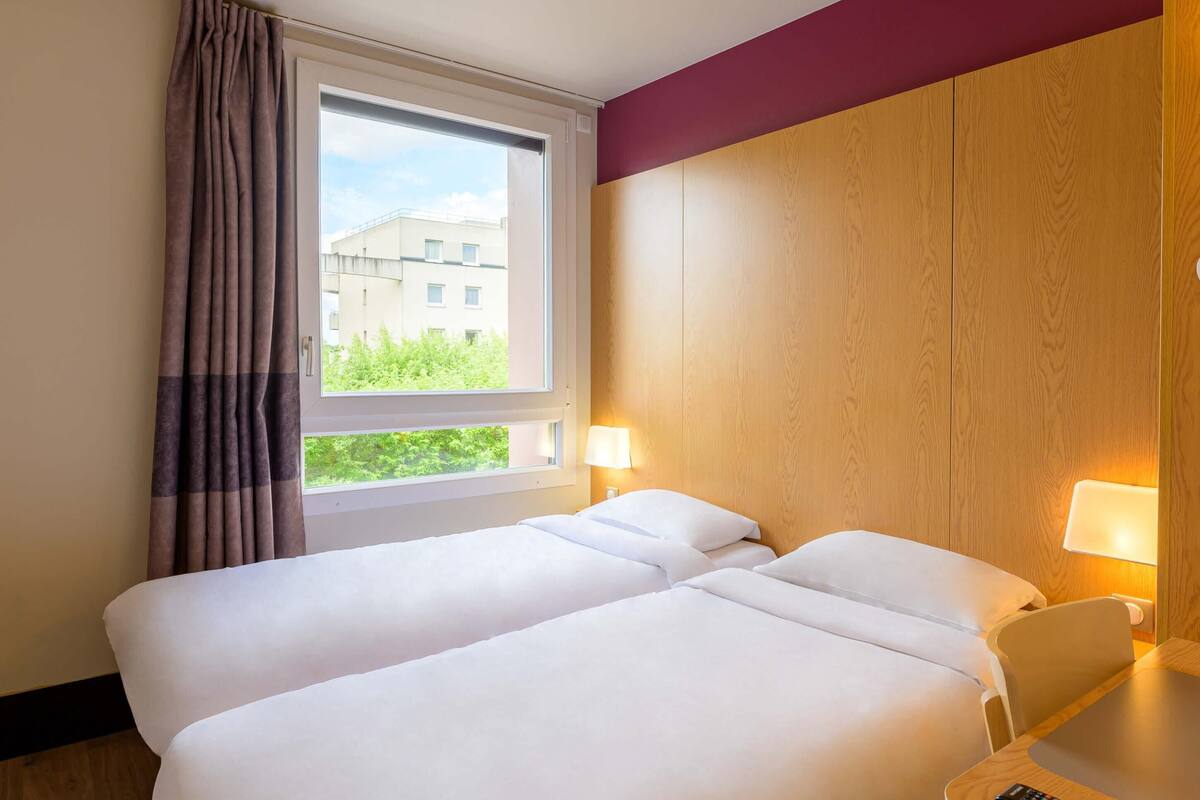 A hotel room with two beds, a window with curtains, a lamp, and a view of greenery outside.