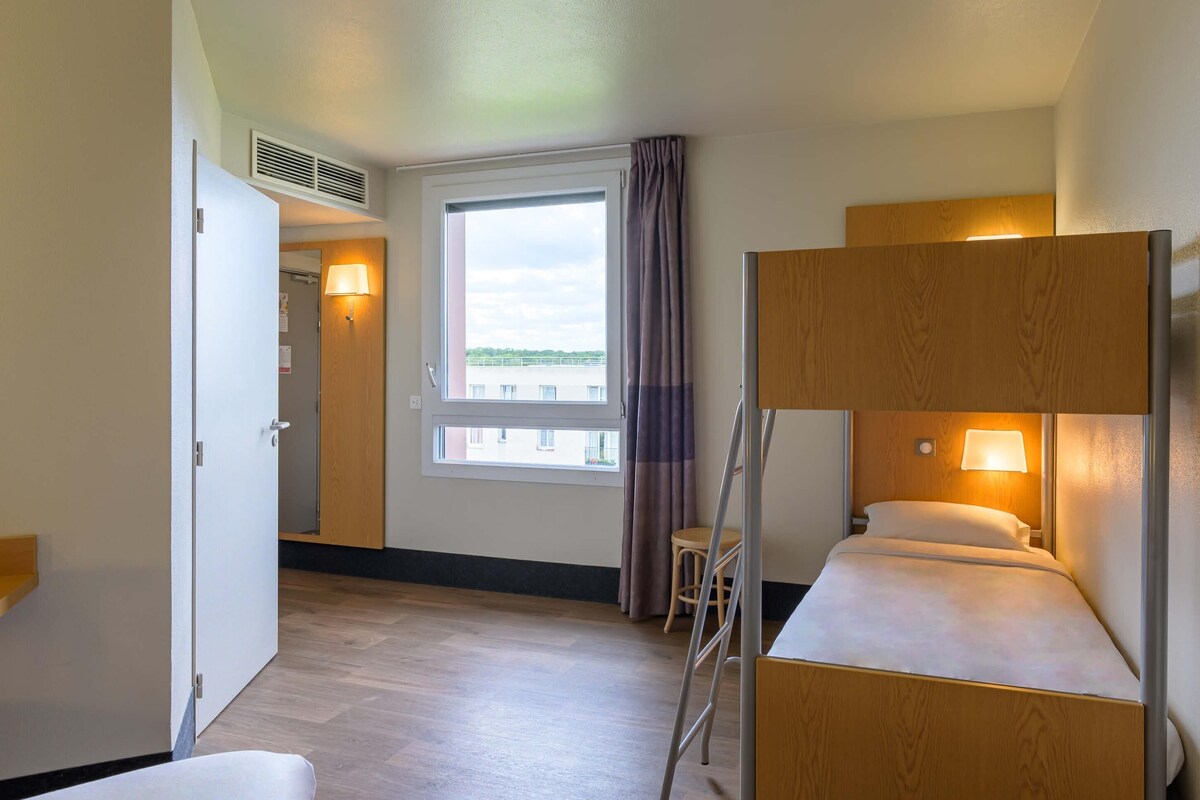 Una habitación de hotel con literas, un escritorio, una silla, una ventana con vistas y una puerta que da a otra habitación.
