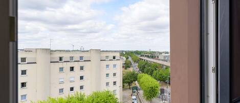 Vista a través de una ventana de un edificio de varios pisos, con cielo despejado, una calle con vehículos y áreas verdes.