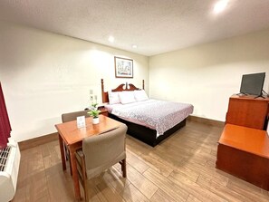 A hotel room with a bed, a desk with a chair, a small table with a plant, and a TV on a stand.