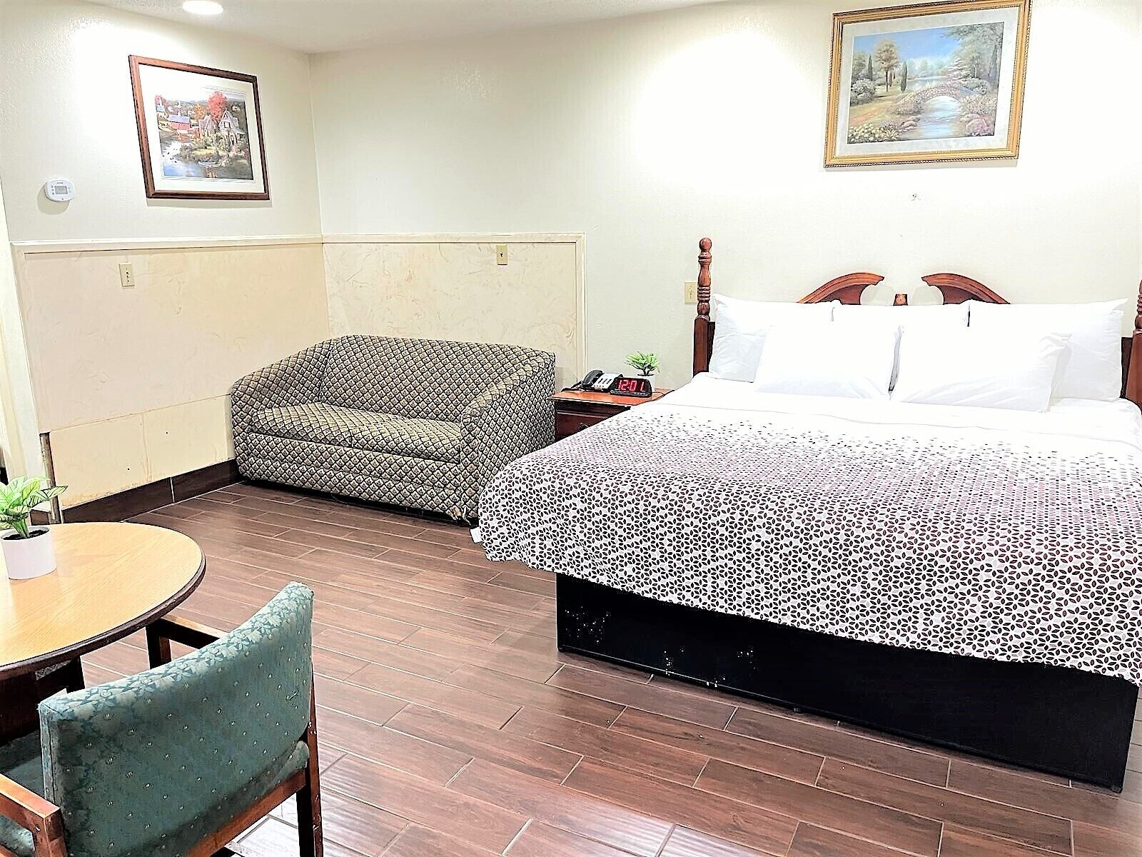 A hotel room with a bed, a chair, a small table, and two paintings on the walls.