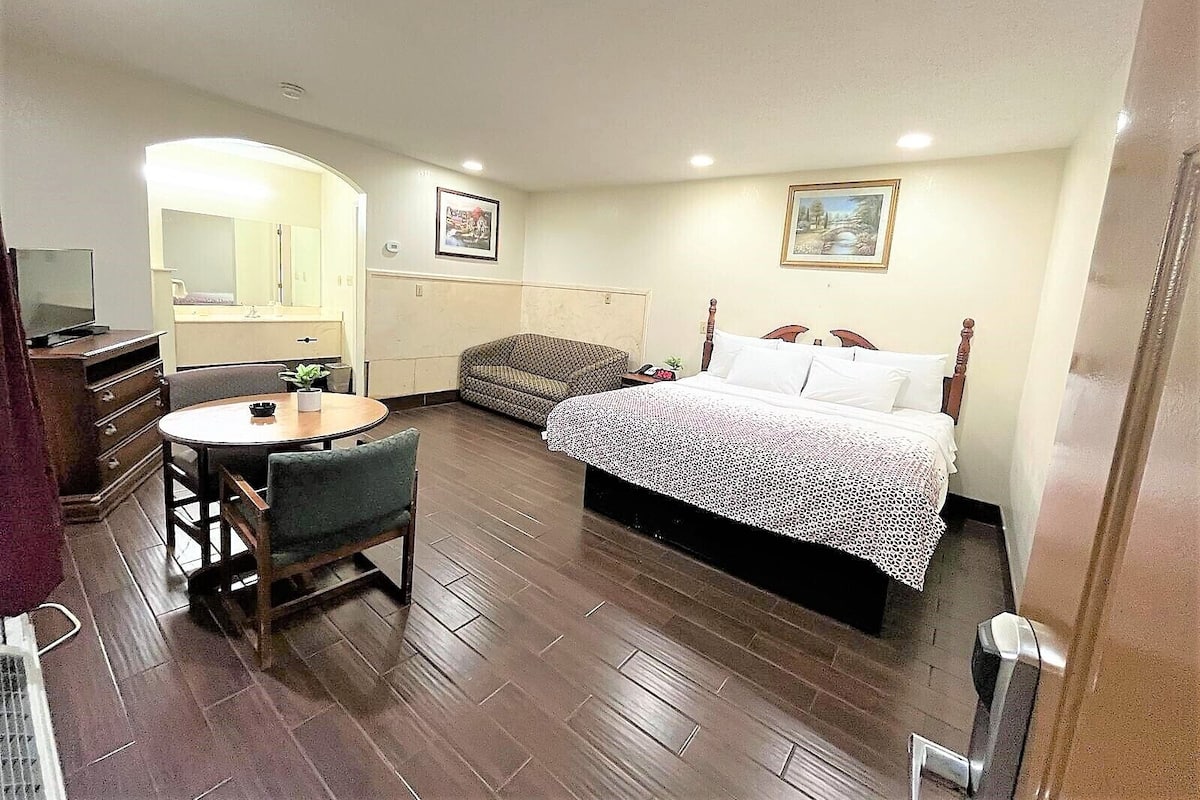 A hotel room with a bed, a small round table with a chair, a sofa, a TV, and a bathroom with a mirror.