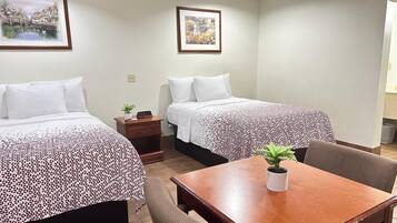 A hotel room with two beds, a small table, and a chair. There are two framed pictures on the wall.