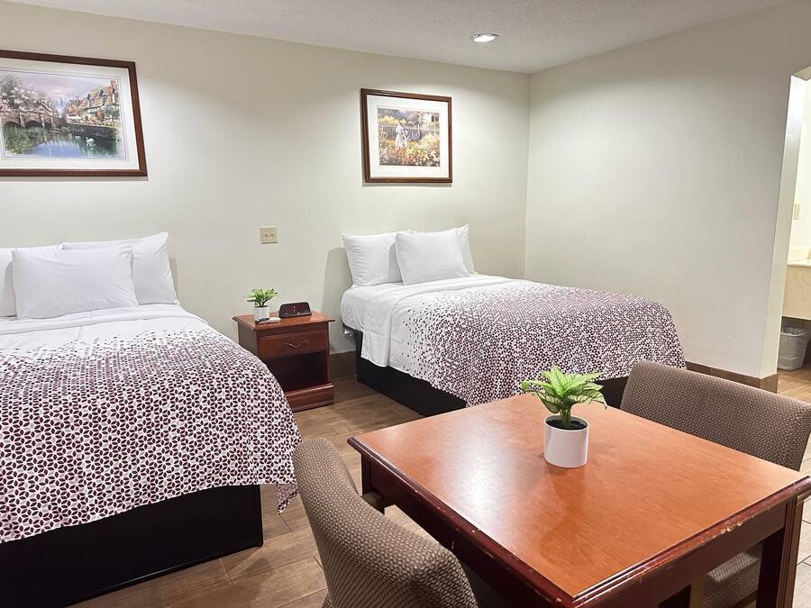 A hotel room with two beds, a small table, and a chair. There are two framed pictures on the wall.